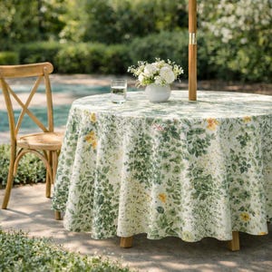 Puede incluir: Una mesa redonda al aire libre con un mantel floral con patrones botánicos verdes y amarillos. A la izquierda hay una silla de madera con un asiento tejido. Un jarrón con flores blancas y un vaso de agua están sobre la mesa. Una sombrilla está en el centro.