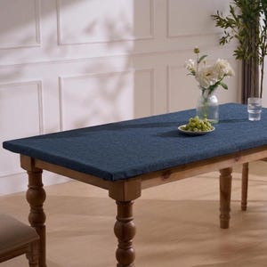 May include: A rectangular wooden table with turned legs is covered by a textured dark blue fabric tablecloth. On the table sits a clear glass vase containing white flowers and a small white plate holding a cluster of green grapes.