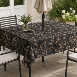 May include: A square outdoor table covered with a dark black tablecloth featuring a repeating pattern of muted brown and beige flowers, leaves, and small birds. A clear glass vase with green foliage sits on the table near a beige patio umbrella.