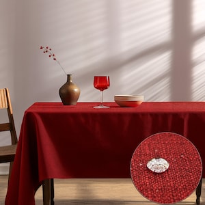 May include: A solid deep red rectangular tablecloth with a textured fabric surface. A circular inset shows a close-up of the fabric with a single clear water droplet resting on top, demonstrating a water-resistant finish on the red material.