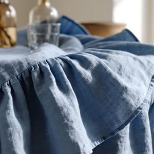 Puede incluir: Un mantel de lino azul claro con un borde con volantes. La tela está drapeada sobre una mesa, con un vaso y botellas en el fondo. El mantel tiene un aspecto suave y texturizado.