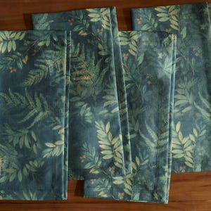 May include: A set of four dark teal napkins with a botanical print. The design features detailed illustrations of green ferns and leaves with small orange berries. The napkins are made of a soft fabric and have a finished hem.