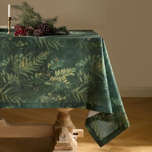 May include: A dark green tablecloth with a botanical fern and leaf pattern. The table is set with a candle holder, pine cones, and red berries. The tablecloth is draped over a wooden table.