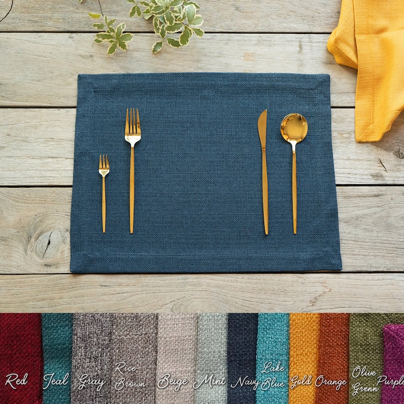 Pu&ograve; includere: Un tovaglietta di lino blu con posate dorate, tra cui una forchetta, un coltello e un cucchiaio. La tovaglietta &egrave; su un tavolo di legno con una variet&agrave; di altre tovagliette di diversi colori visualizzate sotto.