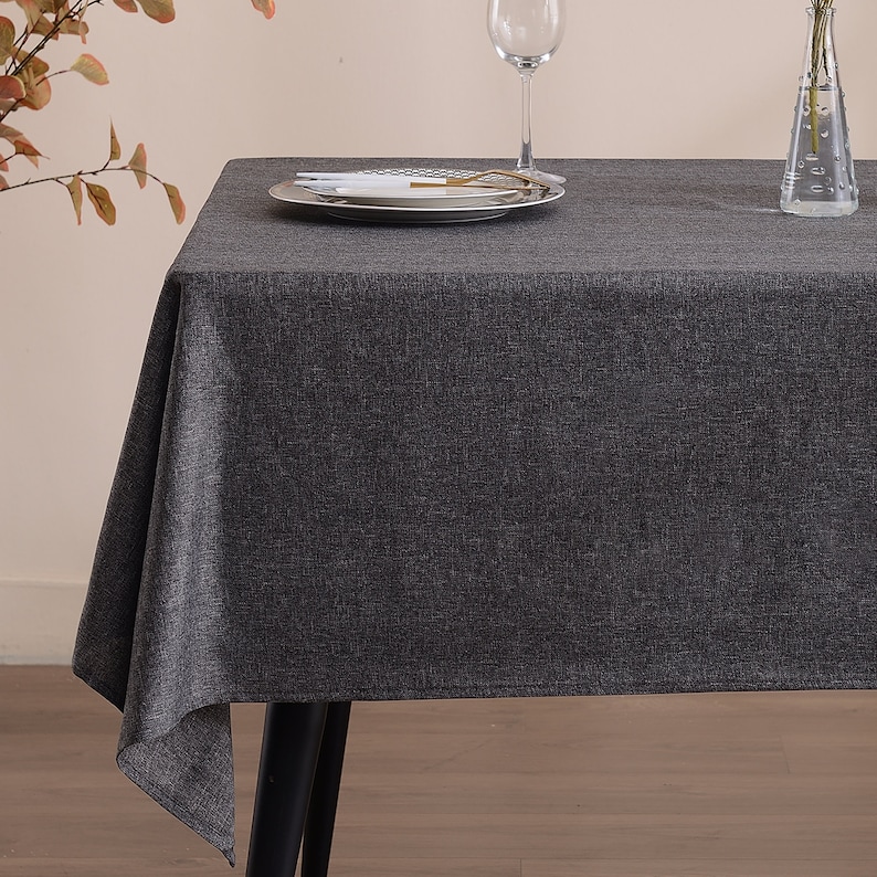 May include: A rectangular dark charcoal gray tablecloth with a visible woven fabric texture draped over a table. The surface features a white plate with cutlery, a clear glass wine goblet, and a clear glass vase containing thin green stems.