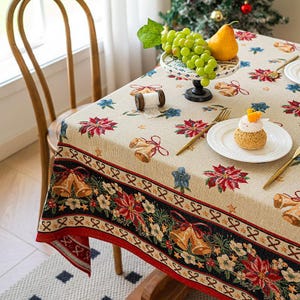 Könnte beinhalten: Beige Weihnachtstischdecke mit einem festlichen Muster aus Glocken, Weihnachtssternen und floralen Designs. Der Rand ist schwarz und rot. Ein Teller mit Gebäck, Trauben und einer Birne steht auf dem Tisch.