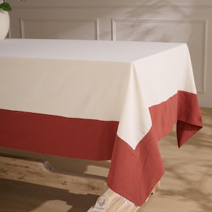May include: A rectangular tablecloth with a two-tone design. The top portion is off-white, and the bottom features a wide band of reddish-brown fabric. The tablecloth is draped over a wooden table, showcasing its texture and color contrast.