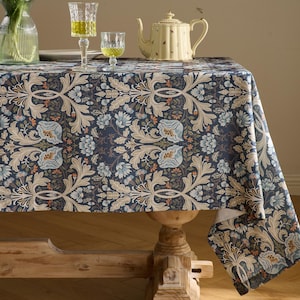 May include: A rectangular tablecloth featuring a dark blue background with a repeating pattern of light beige acanthus leaves, pale blue flowers, and small orange accents. The fabric drapes over a wooden table with a visible pedestal base.