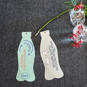 May include: Two bottle-shaped paper tags. The left tag is light green with a blue circular arrow icon, the text Global Recycled Standard, and a GRS logo. The right tag is beige with the text MADE FROM RECYCLED PLASTIC BOTTLES and a graphic.