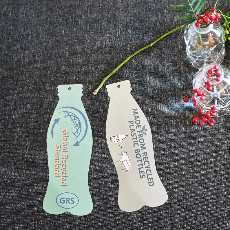 May include: Two bottle-shaped paper tags. The left tag is light green with a blue circular arrow icon, the text Global Recycled Standard, and a GRS logo. The right tag is beige with the text MADE FROM RECYCLED PLASTIC BOTTLES and a graphic.