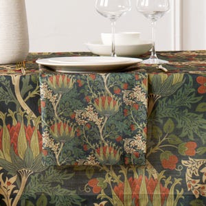 Puede incluir: Una servilleta doblada con un estampado floral en rojo, verde y crema sobre un fondo azul oscuro. La servilleta está colocada sobre un mantel a juego, con platos, copas y un jarrón blancos visibles.