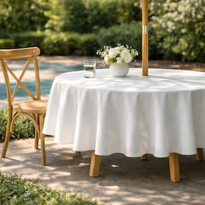 Peut inclure: Une table de salle à manger ronde extérieure avec une nappe blanche, des pieds en bois et un parasol en bois. Une chaise en bois avec une assise en osier est à gauche. Un verre d'eau et un vase blanc avec des fleurs sont sur la table.