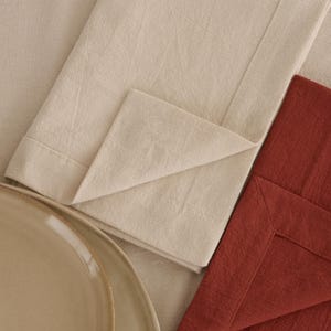 May include: A close-up of a beige folded napkin with a folded corner, a rust-coloured napkin, and a beige plate. The napkins are made of a textured fabric, and the plate has a light brown rim.