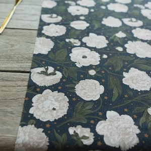 May include: A dark blue floral table runner with large white peony blooms and green leaves. The runner has a repeating pattern of flowers and leaves with small gold stars. Two gold-coloured forks are visible on a wooden surface.