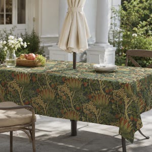 May include: A rectangular tablecloth featuring a dark green background with a repeating botanical pattern of large orange and green artichoke-like flowers, winding stems, and leaves, draped over an outdoor table with a central umbrella pole.