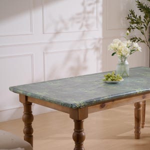 May include: A rectangular wooden table featuring turned legs and a dark green tablecloth with a botanical leaf pattern. A clear glass vase containing white flowers and a small white bowl filled with green grapes sit on the table surface.