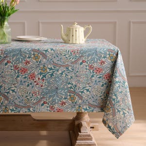 May include: A rectangular tablecloth featuring a dense, intricate botanical pattern of scrolling blue leaves, pink flowers, and yellow blossoms on a cream background. The fabric drapes over a wooden table, with a cream-colored teapot placed on top.