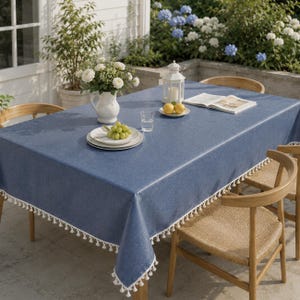 May include: A rectangular blue tablecloth with a textured finish and white tassel fringe trim along the edges. The tablecloth is draped over a table, featuring a white ceramic pitcher with flowers, a plate of green grapes, and a white lantern.