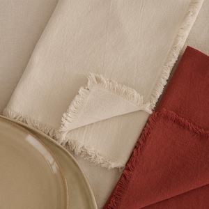 Può includere: Vista dall'alto di tovaglioli di lino piegati, color crema e ruggine, con bordi sfrangiati. Una porzione di un piatto in ceramica beige è visibile in basso a sinistra. I tovaglioli sono disposti su una superficie dai toni neutri.