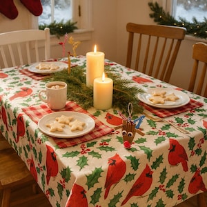 Könnte beinhalten: Ein festlicher Weihnachtstisch mit einer Tischdecke mit roten Kardinälen und grünem Stechpalmenmuster. Der Tisch ist mit Tellern mit sternförmigen Keksen, Tassen mit heißer Schokolade und brennenden Kerzen gedeckt. Ein Holz-Rentier-Ornament sorgt für eine dekorative Note.