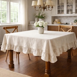 May include: A square, cream-colored linen tablecloth with a ruffled edge, draped over a wooden table. A white vase with white flowers sits in the center. The room has a chandelier and wooden chairs.