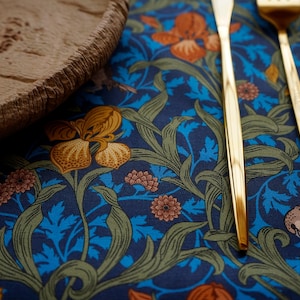 May include: Two gold-colored flatware pieces, a knife and a fork, are laid on a blue and green floral patterned tablecloth. The tablecloth has a repeating pattern of flowers, leaves, and birds.