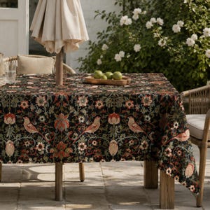 Könnte beinhalten: Eine rechteckige Tischdecke mit schwarzem Hintergrund und einem sich wiederholenden Muster aus orangefarbenen Vögeln, roten Blumen und grünem Blattwerk. Der Stoff liegt auf einem Tisch mit einem zentralen Schirmstock und einer Holzschale.