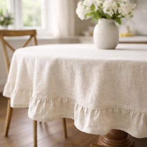 May include: A cream-coloured, round tablecloth with a ruffled edge. The tablecloth is draped over a wooden table, with a vase of white flowers in the centre. The table is set in a bright, airy room.
