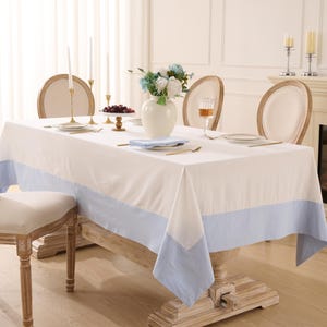 May include: A rectangular white tablecloth with a light blue border covers a wooden dining table. The table is set with plates, silverware, and a vase of flowers. Two chairs are visible, and the room has a bright, airy feel.