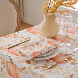 May include: A white tablecloth featuring a pumpkin and leaf design, ready for an autumn meal. The table is set with plates, napkins, and gold cutlery. Two wine glasses and a vase with orange branches are also present.