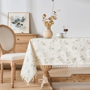 Pode incluir: Uma cena de sala de jantar com uma toalha de mesa floral creme com acabamento de borlas. A mesa está posta com um vaso de flores secas, uma chávena de chá e uma taça de vinho. Duas cadeiras de madeira com encostos ovais e uma cómoda de madeira também são visíveis.