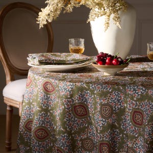May include: A round tablecloth with a green, red, and white floral pattern. The table is set with white plates, green patterned napkins, and a bowl of cherries. A white vase with dried flowers and a wooden chair are in the background.