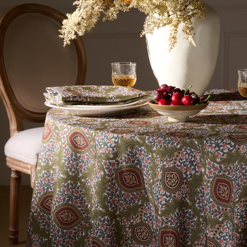 May include: A round tablecloth with a green, red, and white floral pattern. The table is set with white plates, green patterned napkins, and a bowl of cherries. A white vase with dried flowers and a wooden chair are in the background.