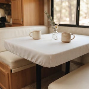 Könnte beinhalten: Ein rechteckiger Tisch mit einem passgenauen, hellbeigen Stoffbezug. Auf der Oberfläche stehen zwei gesprenkelte, helle Keramiktassen sowie eine durchsichtige Glasvase mit einem kleinen Arrangement aus Eukalyptusblättern und weißen Gänseblümchen.