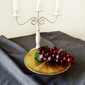 May include: A white metal candelabra with three arms and a distressed finish, positioned behind a circular gold-toned metal tray. A cluster of artificial dark red and purple grapes with green leaves rests on the surface of the gold tray.