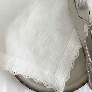 May include: A white linen napkin with an embroidered letter "R" in the center. The napkin has a delicate lace trim around the edges and is placed on a plate with silverware.