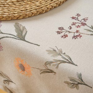 Puede incluir: Primer plano de un mantel beige con motivos florales con diseños de flores y hojas de estilo acuarela en tonos rosa, naranja y verde. Un salvamanteles de paja tejida está en la esquina superior izquierda.