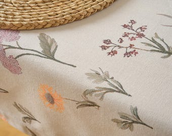 May include: Close-up of a beige floral tablecloth with watercolor-style flower and leaf designs in shades of pink, orange, and green. A woven straw placemat is in the upper left corner. The fabric has a textured appearance.