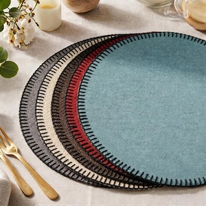 May include: A stack of round placemats in various colors: gray, cream, brown, red, and teal. Each placemat has a black stitched border. Gold forks are visible in the lower left corner. The placemats are on a light-colored surface.