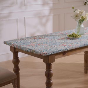 May include: A wooden dining table with a floral patterned tablecloth in shades of blue, red, and yellow. The table has turned legs and is set with a vase of white flowers and a plate of green grapes.