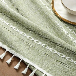 Könnte beinhalten: Ein grüner Tischläufer mit weißen Stickereien und Fransen. Der Läufer hat eine gewebte Textur und einen dezenten Glanz.