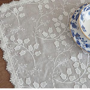 Puede incluir: Mantel blanco bordado con diseño floral y bordes festoneados. Un plato y un cuenco con estampado floral azul y blanco se encuentran sobre el mantel, creando una estética delicada y elegante.
