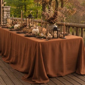 May include: A solid rust-colored tablecloth draped over a long rectangular table. The table is set with white pumpkins, dark metal lanterns, tall taper candles, textured glassware, and a large vase filled with dried foliage and branches.