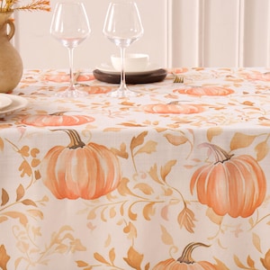May include: A white tablecloth with a repeating pattern of orange pumpkins and autumn leaves. The pumpkins have brown stems and are shaded with orange and peach tones. The tablecloth is set with wine glasses and plates.