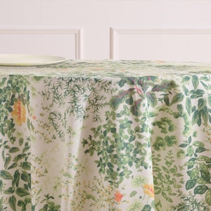 May include: A round tablecloth with a botanical print featuring green leaves and yellow flowers on a cream background. The design includes various leaf shapes and sizes, creating a natural, garden-inspired aesthetic. A light-colored plate is visible in the upper left corner.