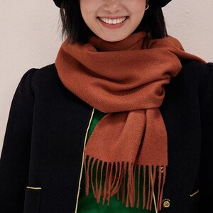 May include: A rust-colored scarf with fringe detail is draped over a black coat. The scarf is made of a soft material and is wrapped around the neck. The coat has gold trim and a green shirt is visible underneath.