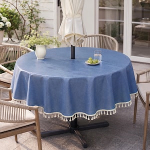 May include: A round blue tablecloth with a textured finish and a cream-colored fringe trim along the edge. The tablecloth is draped over a circular outdoor table that features a central pole for an umbrella, a small white vase, and a glass.