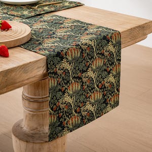 Puede incluir: Un camino de mesa verde y negro con un patrón floral con flores rojas y naranjas. El camino de mesa está sobre una mesa de madera con un tazón de madera y fresas rojas.