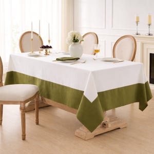 Puede incluir: Un mantel rectangular blanco con un borde verde oliva, colocado sobre una mesa de madera. La mesa está puesta con platos, servilletas y cubiertos. Se ven dos sillas y un jarrón con flores blancas en el centro.