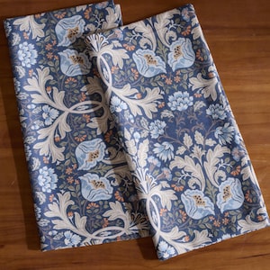 Pode incluir: Duas toalhas de tecido dobradas com padrão floral azul. As toalhas apresentam um padrão repetido de flores azul claro, folhas bege e pequenos detalhes laranja sobre um fundo azul escuro. As toalhas são feitas de um material macio, provavelmente algodão.